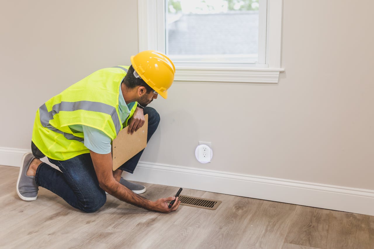 A construction worker in a hard hat inspects a floor vent indoors, ensuring quality and safety standards.